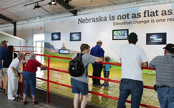People standing a railing overlooking a giant map of Nebraska on the floor.